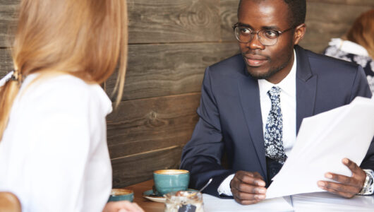 Recruiting and human resource concept. African businessman interviewing Caucasian female candidate for a secretary position, looking and listening to her attentively while sitting at a coffee shop